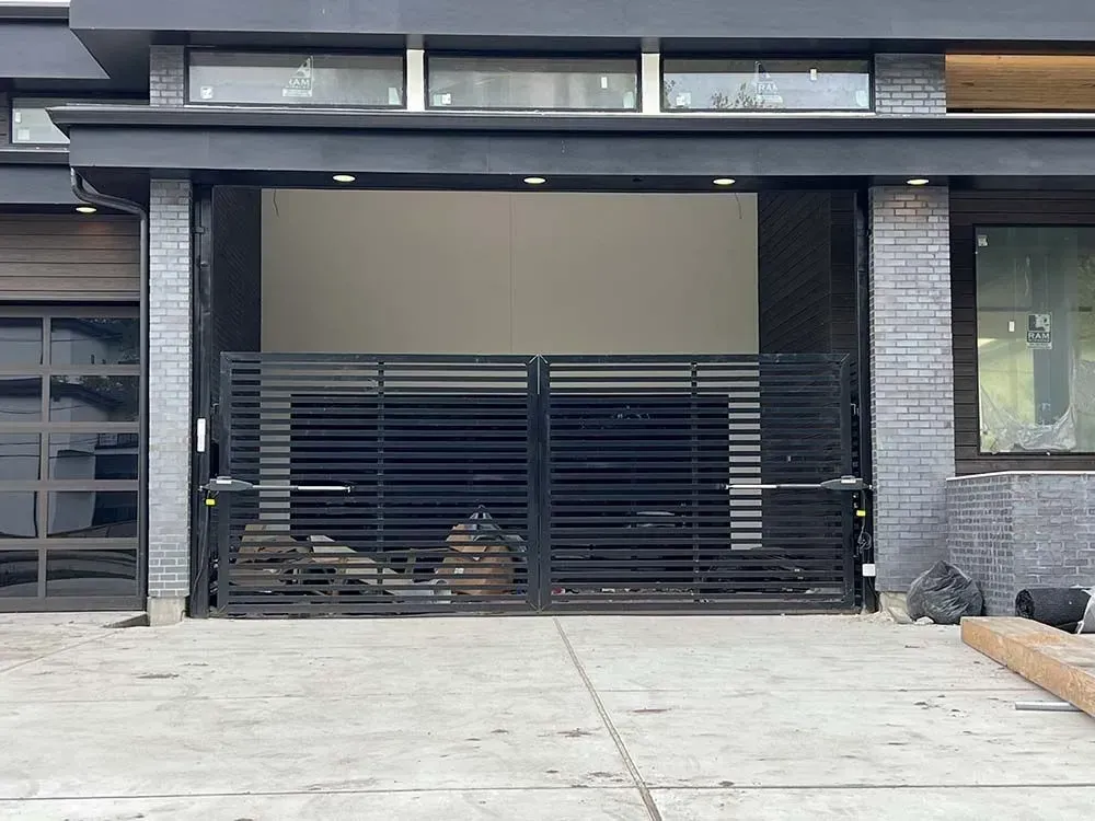 Black metal driveway gates, open, in front of a modern home with glass garage door and brick accents.