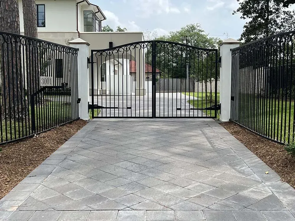 Black wrought-iron driveway gate, brick pavers, and matching fence leading to a white house.