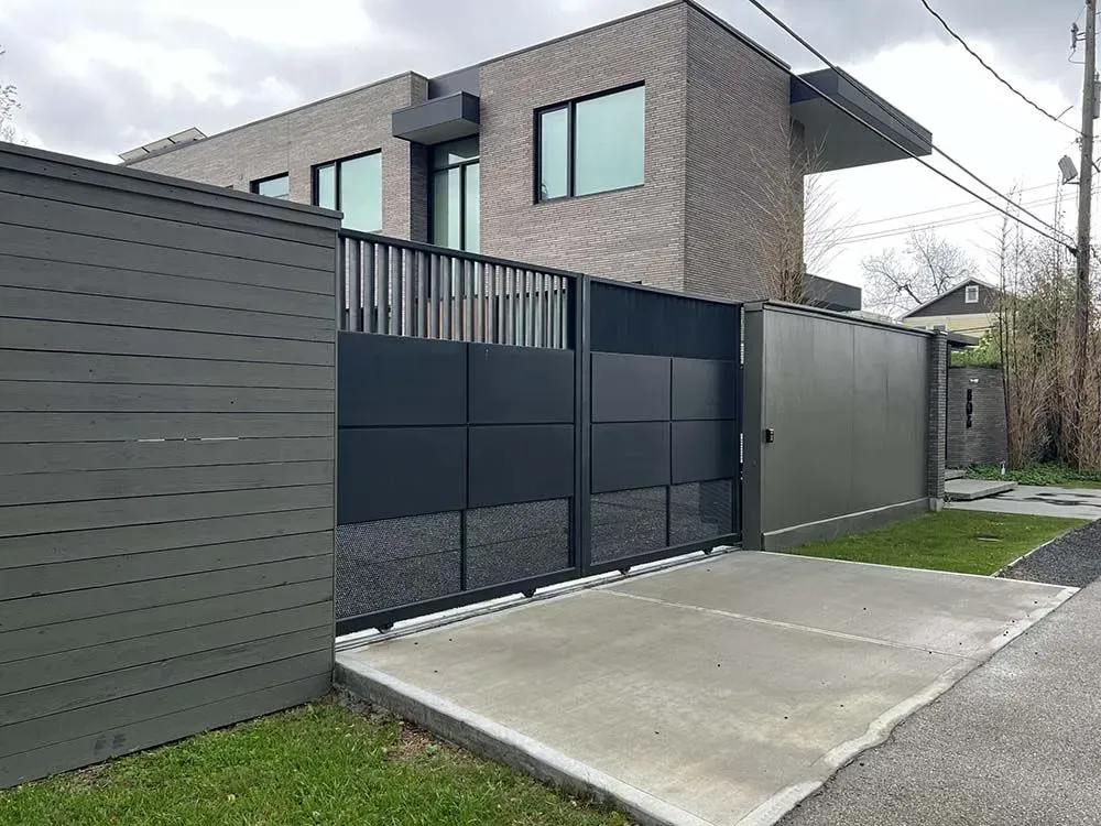 Modern home with gray brick facade, enclosed by a black metal gate and fence. Concrete driveway.