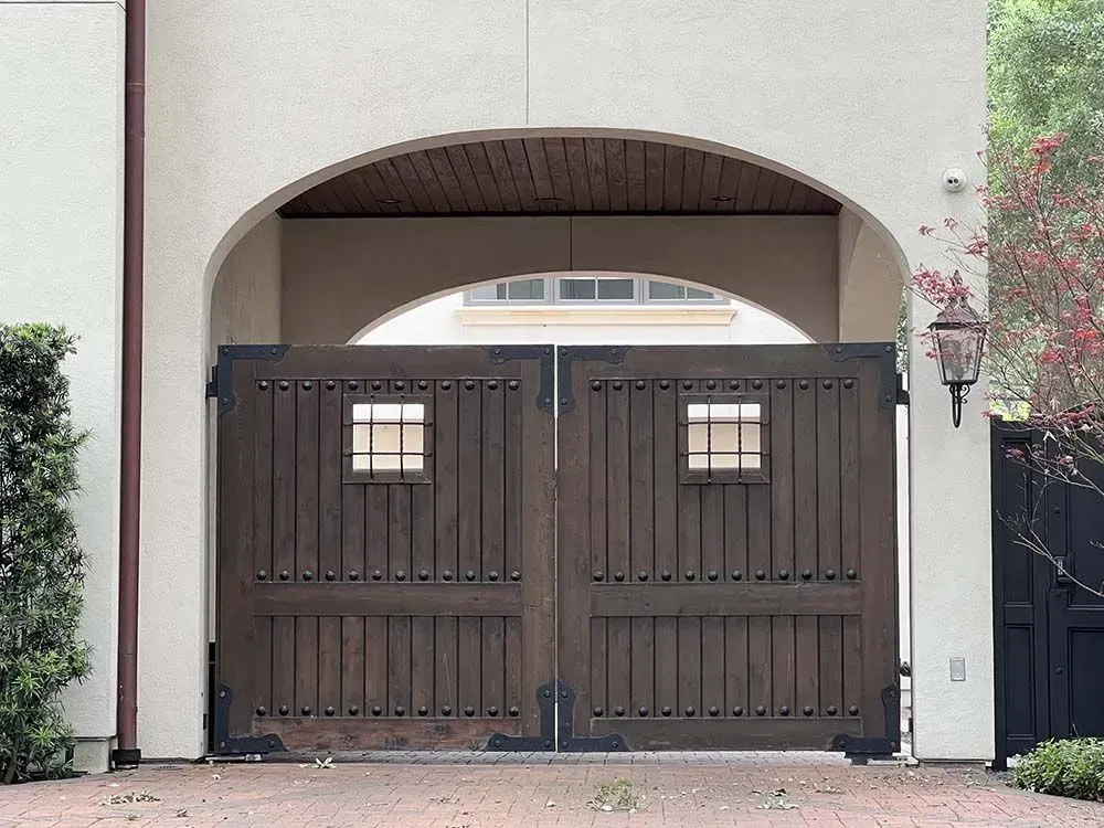Wooden gate under an arched entryway, dark brown with small square windows. Brick driveway and cream-colored walls.