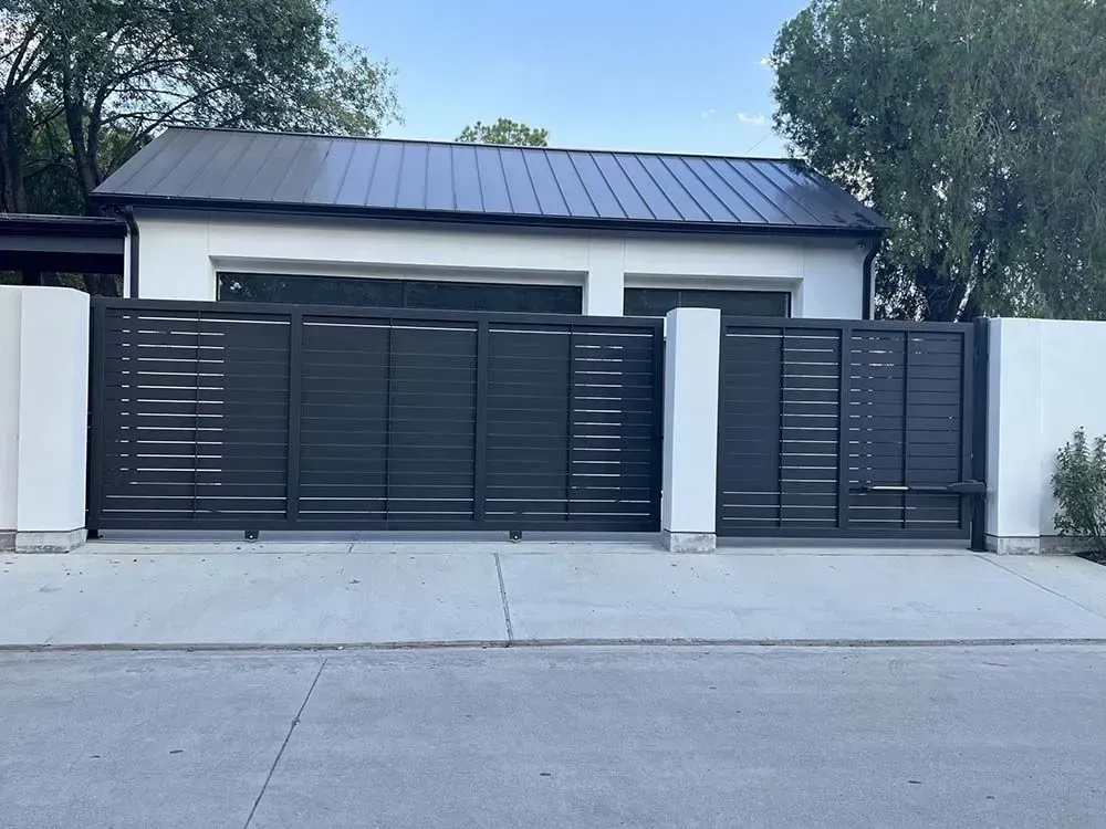 Dark gray horizontal slat gate in front of a white building with black roof. Concrete driveway.