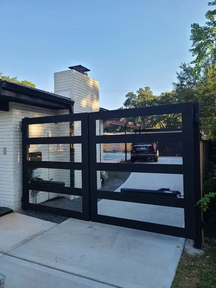 Black metal and glass gate, in front of a white brick house, on a concrete surface.