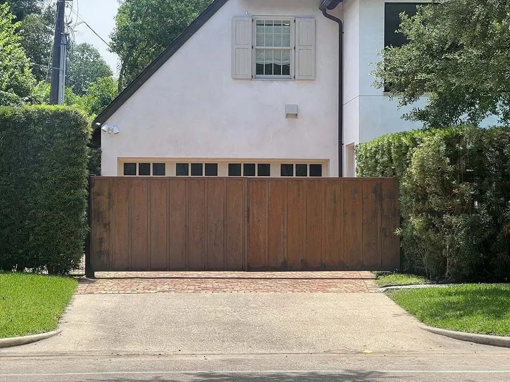 Brown wooden gate blocking a driveway leading to a white house with a gabled roof.