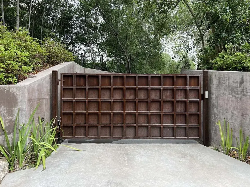 Brown grid gate set in concrete walls. Lush green foliage surrounds.