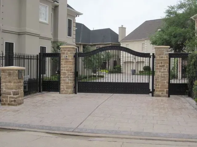 Black wrought iron driveway gates with stone pillars and a patterned concrete driveway.