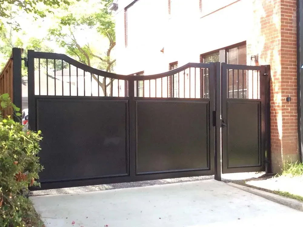 Black metal driveway gate with vertical bars and solid panels, against a white building.