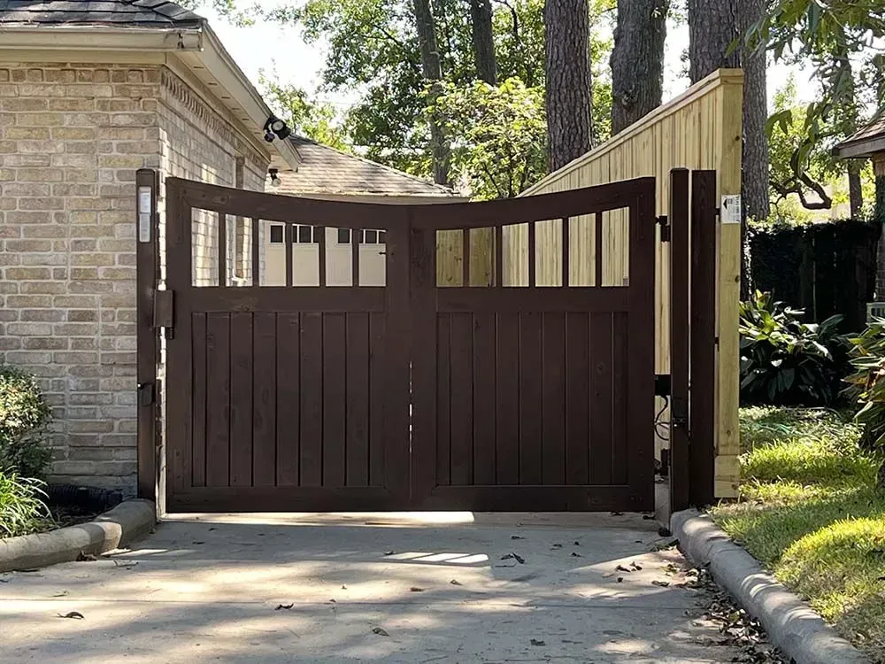 Brown wooden gate in front of a driveway leading to a house. Brick building to the left, fence to the right.
