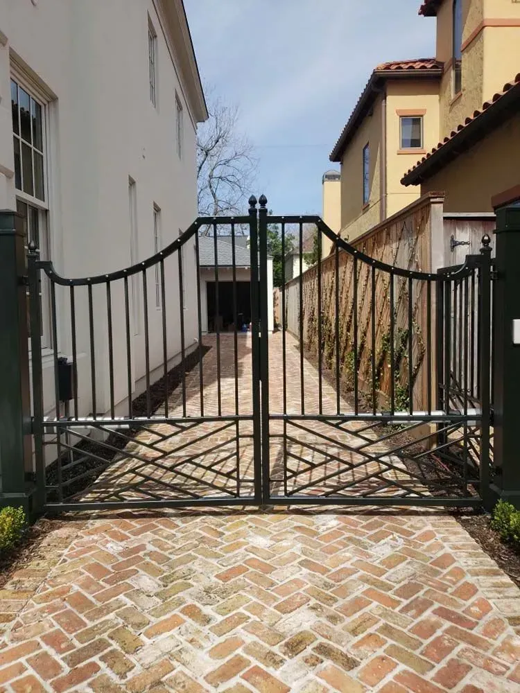 Black wrought iron driveway gates on brick pavers between two buildings.