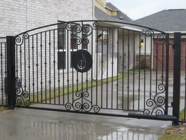 Black wrought-iron driveway gate with decorative swirls and a circular emblem in front of a house.