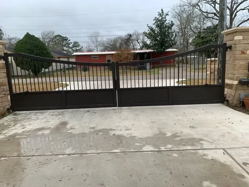 Dark brown metal driveway gates opening onto a concrete driveway, set between stone columns; house visible in the background.