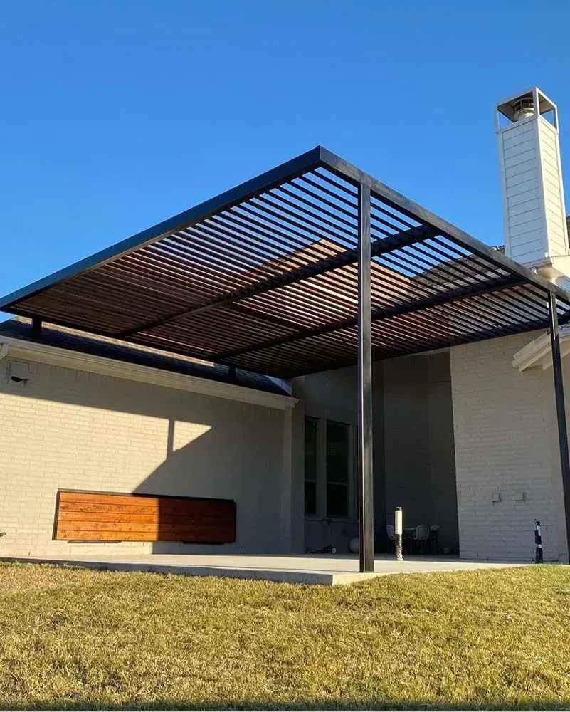 Black metal pergola over a patio, attached to a light brick house with a wooden bench.
