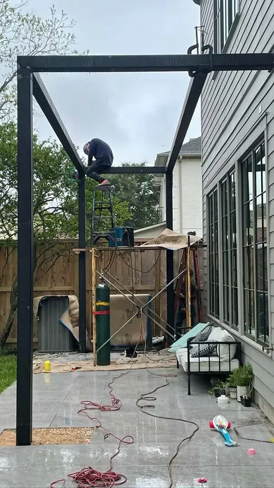 A person welds a metal frame on a patio. Tools, a gas tank, and a wood fence are nearby.