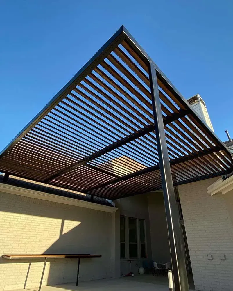Black metal pergola with brown slats over a patio, against a brick wall. Sunny day.