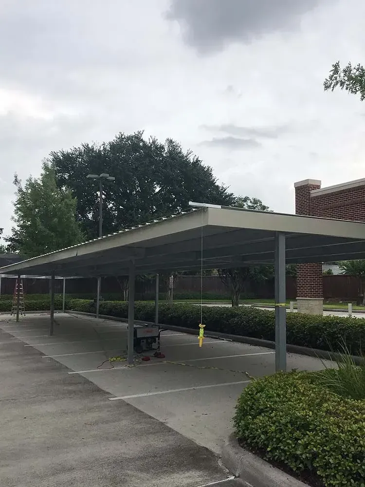 Covered parking area with metal supports, gray roof, concrete surface, and green bushes.