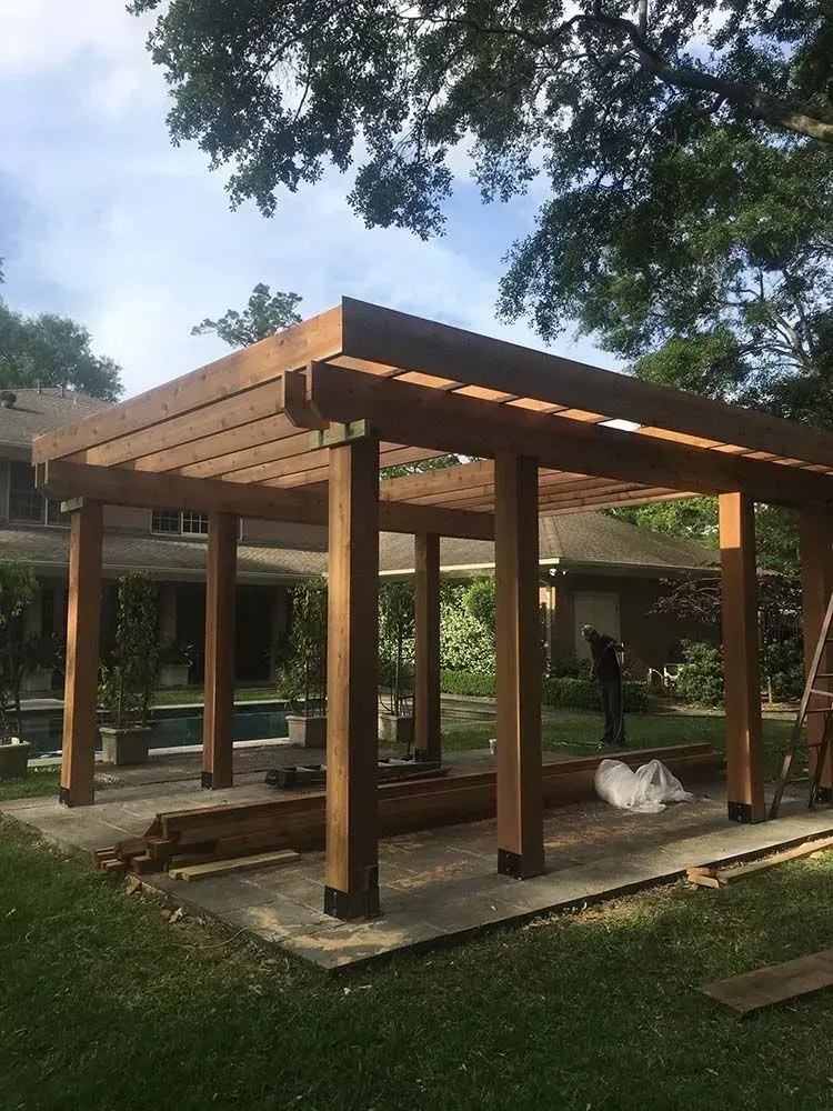 Wooden pergola under construction in a backyard, with a person working in the background.