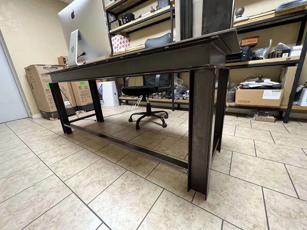 Steel-framed desk with a computer monitor, office chair, and shelves in the background, set on a tiled floor.