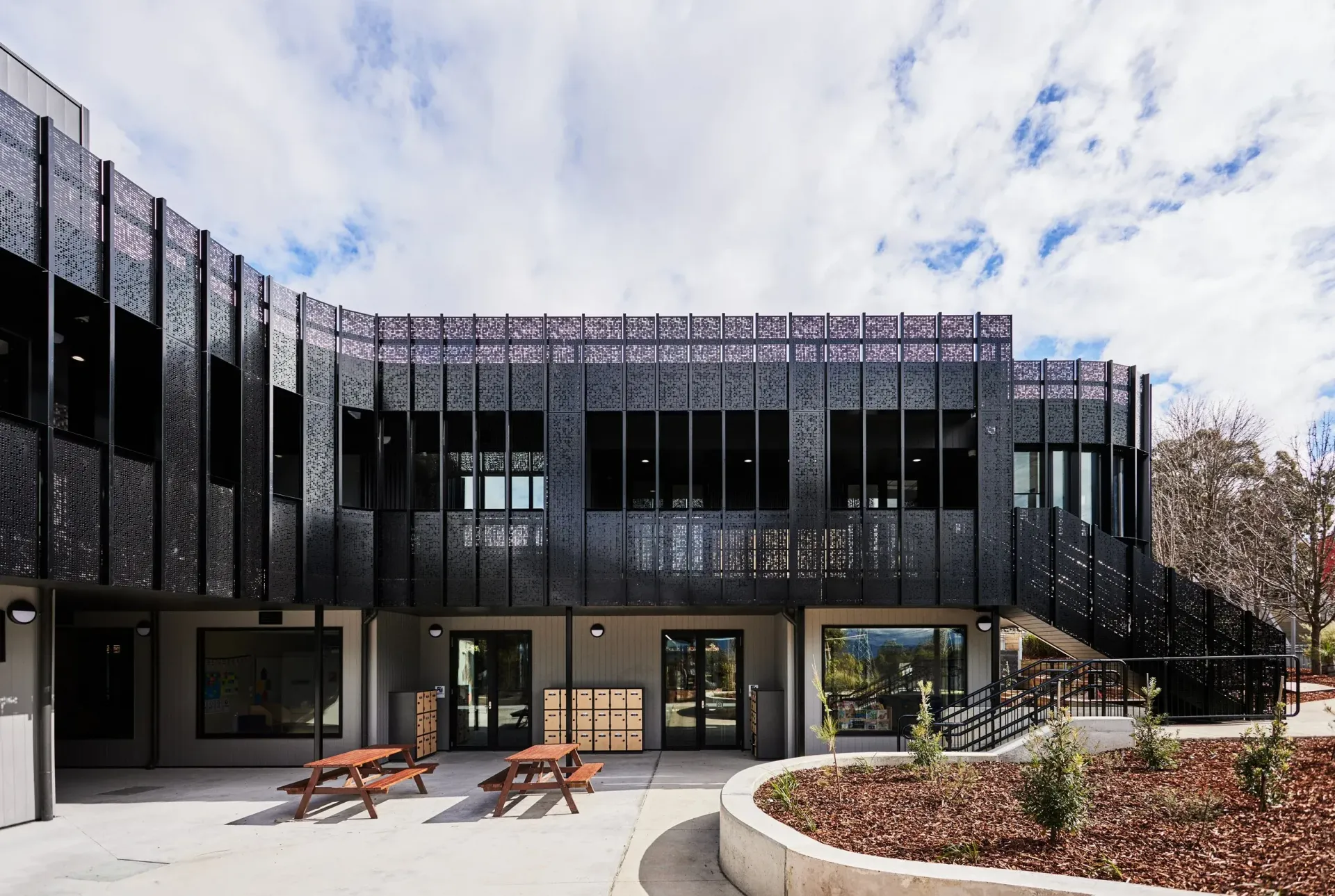 Modern building with black perforated facade, outdoor seating, and cloudy sky.