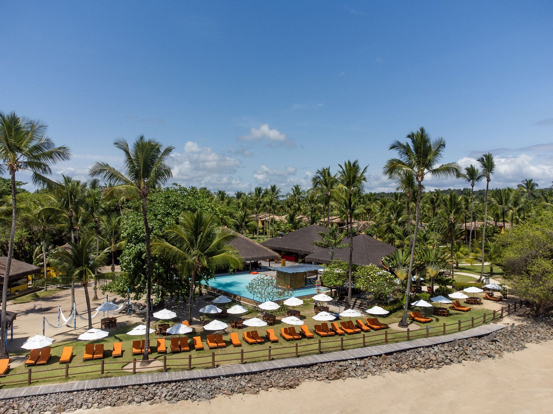 Resort à beira da piscina com espreguiçadeiras laranja, guarda-sóis e palmeiras sob um céu azul.