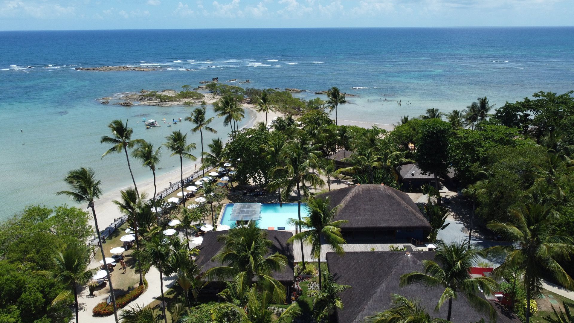 Vista aérea de um resort tropical com praia, piscina, palmeiras e oceano sob um céu azul.
