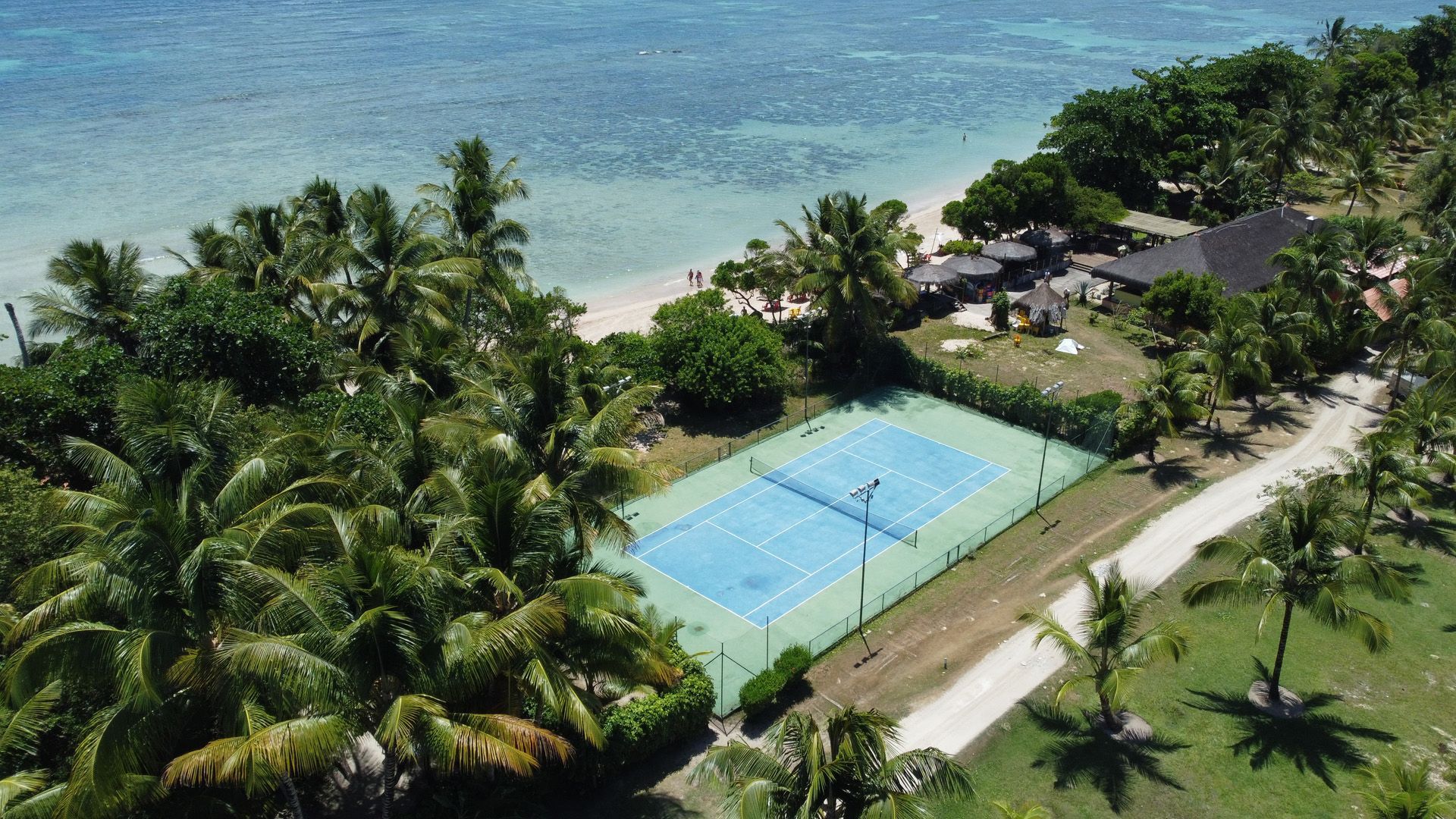 Vista aérea de uma quadra de tênis ao lado de uma praia com palmeiras e um prédio.