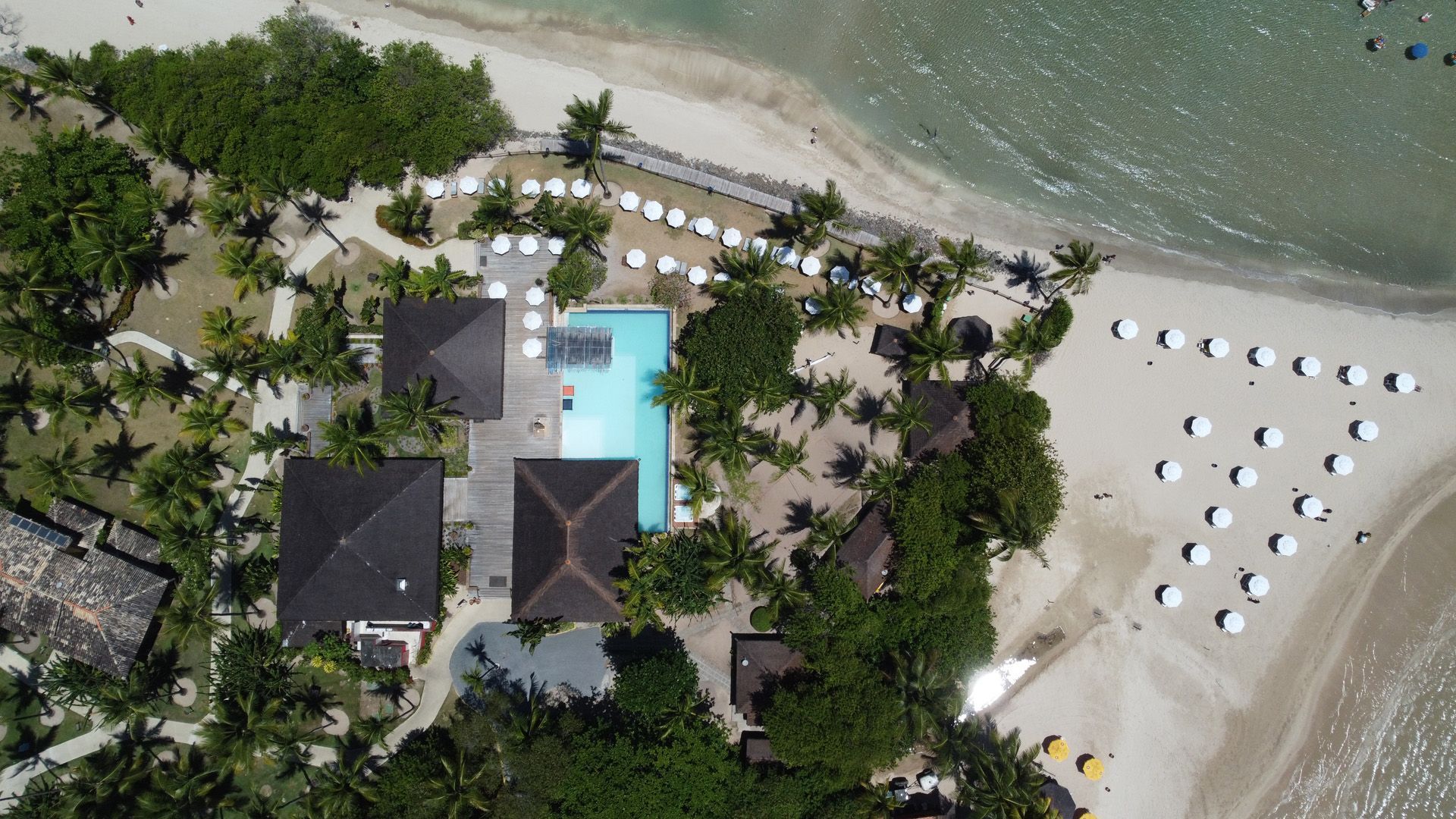 Vista aérea de um resort com piscina, edifícios, praia e guarda-sóis perto do oceano.