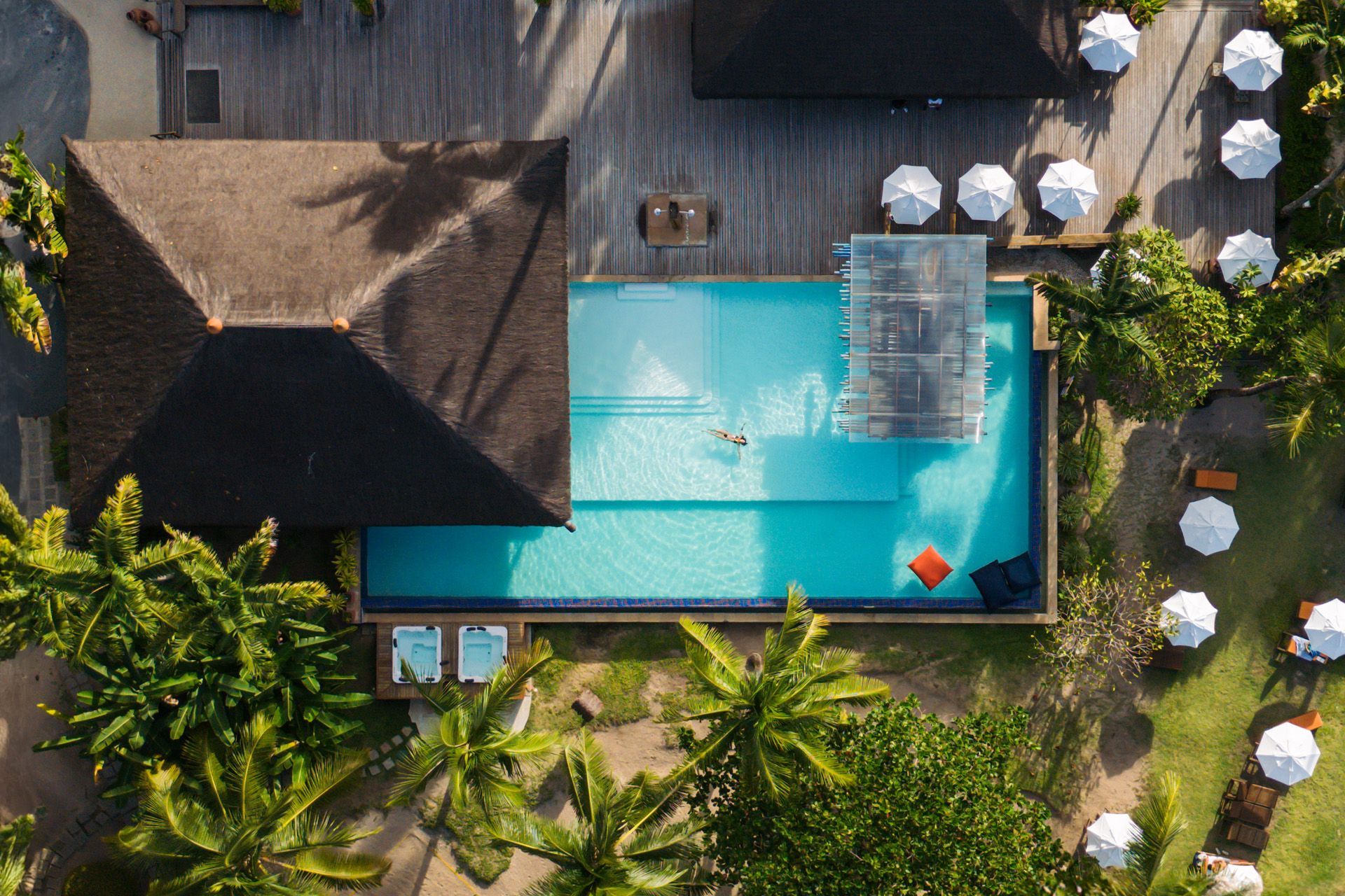 Vista aérea de uma piscina retangular com uma pessoa nadando perto de uma estrutura de madeira, cercada por construções com telhados de palha e guarda-sóis.