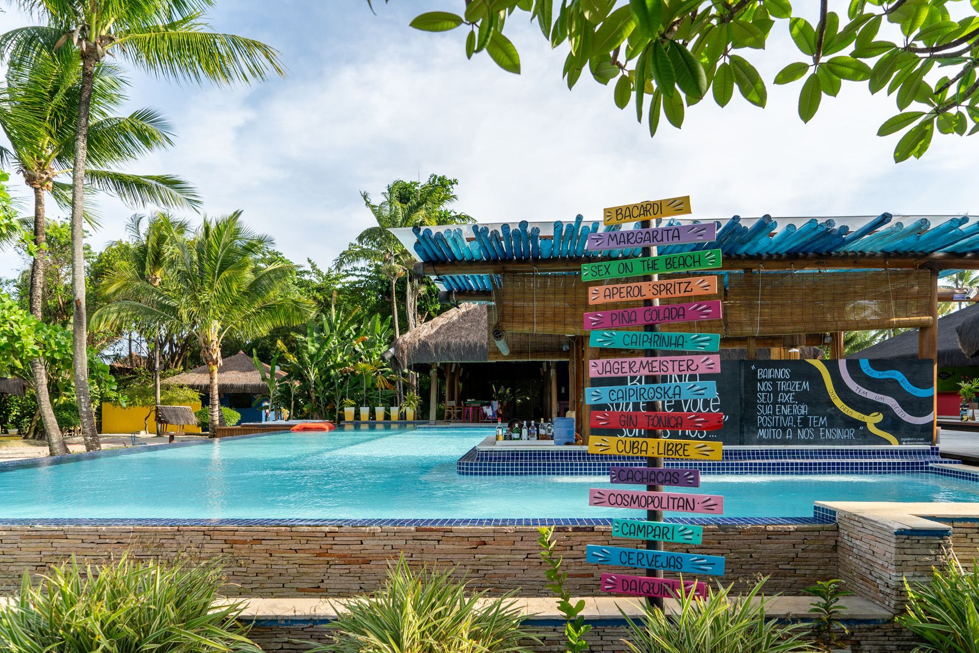 Mulher de biquíni sentada à beira de uma piscina, sombreada por um bar azul e de bambu, sob um céu ensolarado.