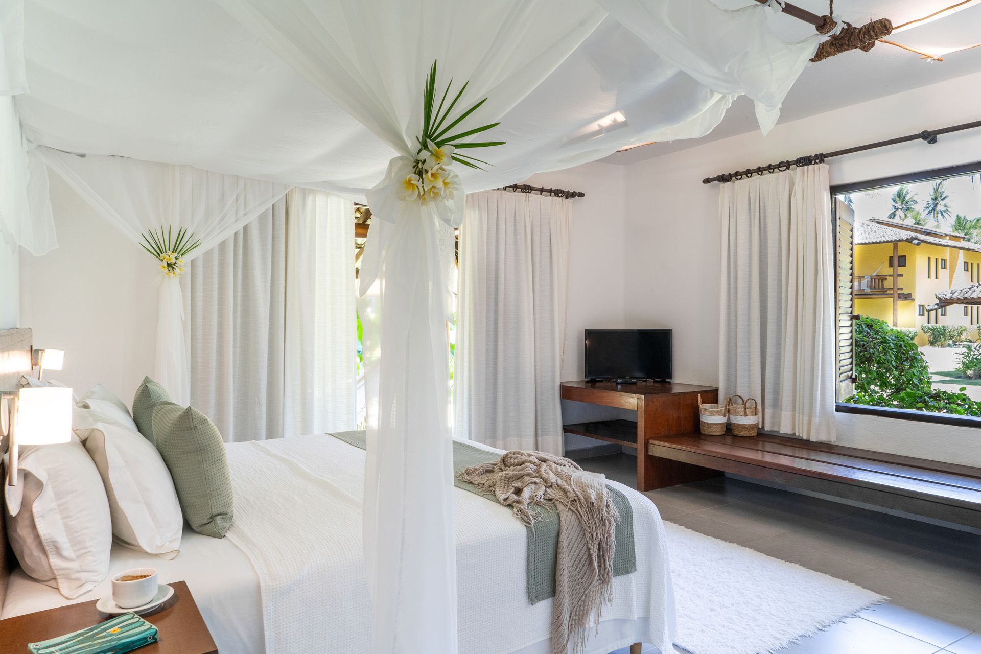 Quarto com cama de dossel, almofadas verde-claras, decorações florais e arte na parede.