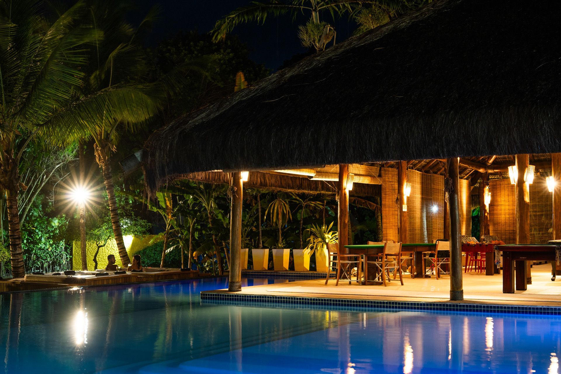 Cena noturna de piscina com bar de teto de palha, iluminada por luzes aconchegantes e palmeiras.