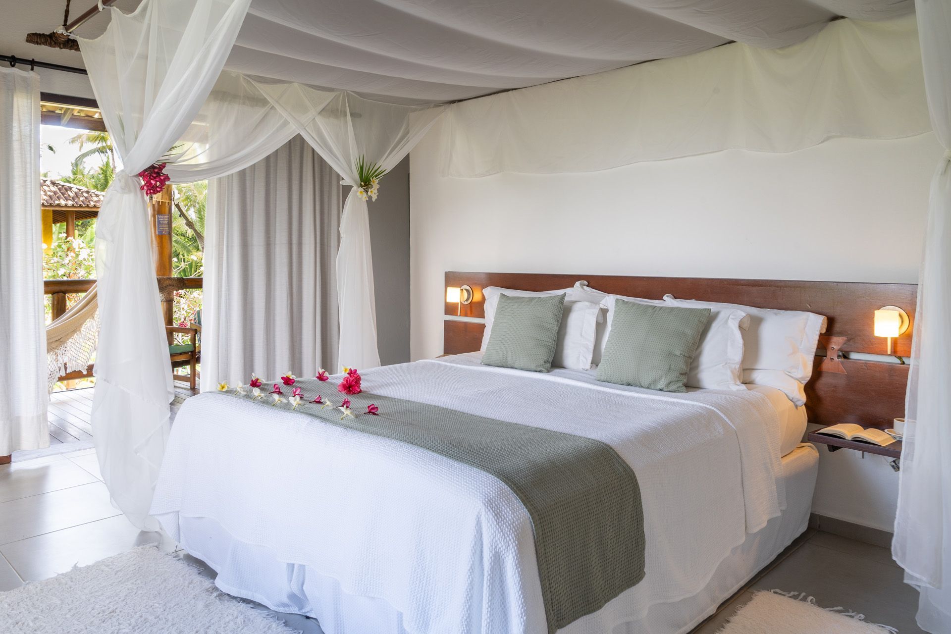 Quarto com cama de dossel, roupa de cama branca, almofadas verdes e detalhes florais. Dá acesso a uma varanda.