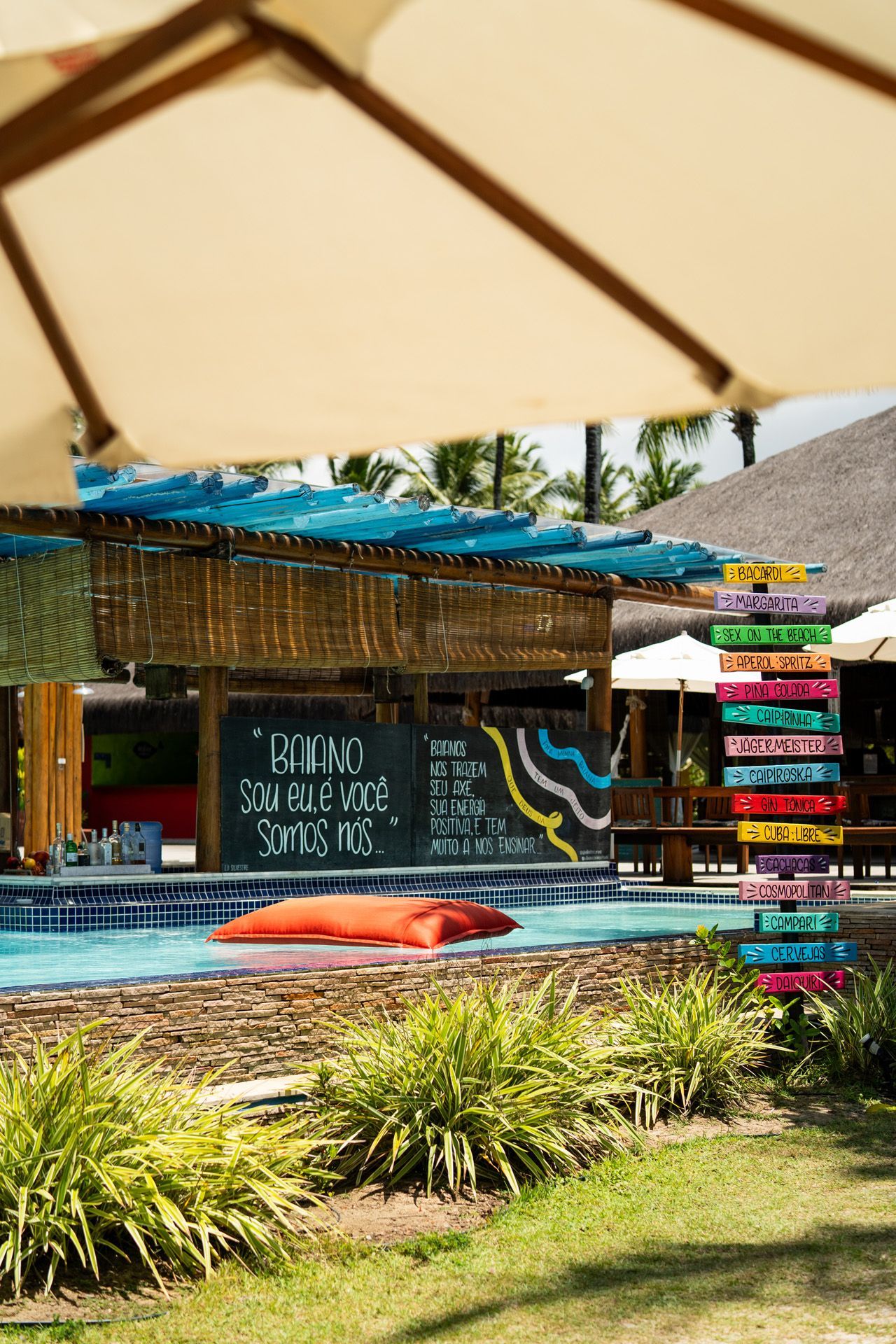 Bar à beira da piscina com teto de palha, pufes flutuantes e placas coloridas empilhadas.