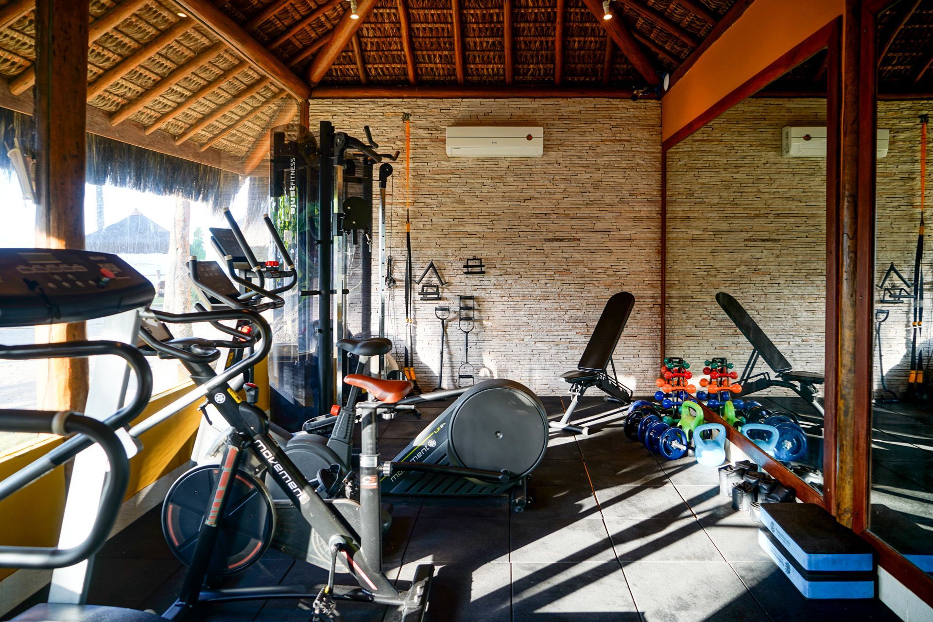 Academia com equipamentos de ginástica dentro de uma estrutura com teto de madeira, apresentando uma parede de tijolos e um grande espelho.