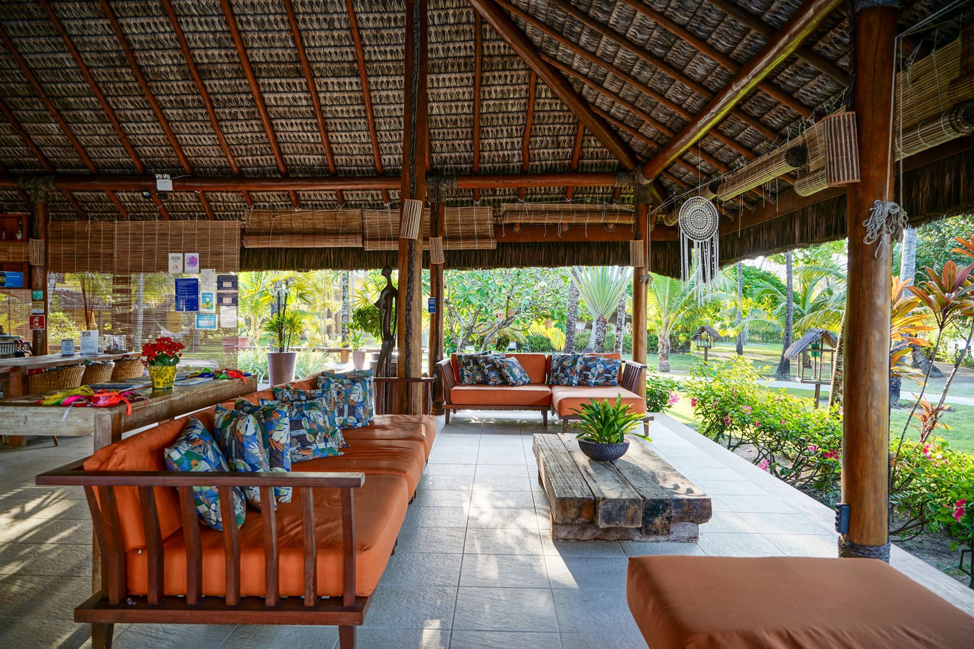 Área de estar externa com móveis de madeira, teto de palha e vegetação exuberante.