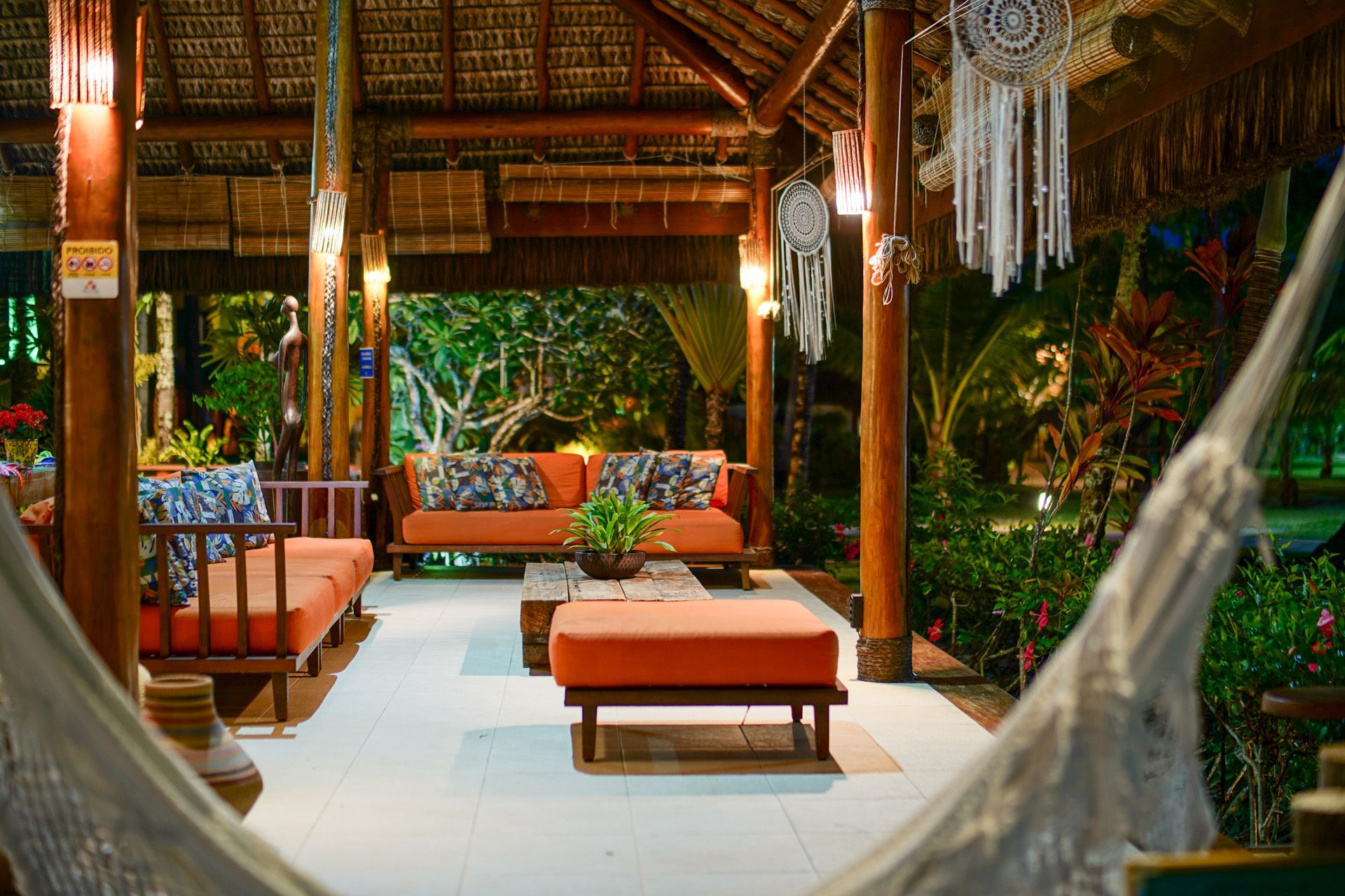 Espaço de convivência aconchegante e ao ar livre com móveis de madeira, almofadas laranja e apanhadores de sonhos; iluminado por luzes quentes com folhagem exuberante ao fundo.