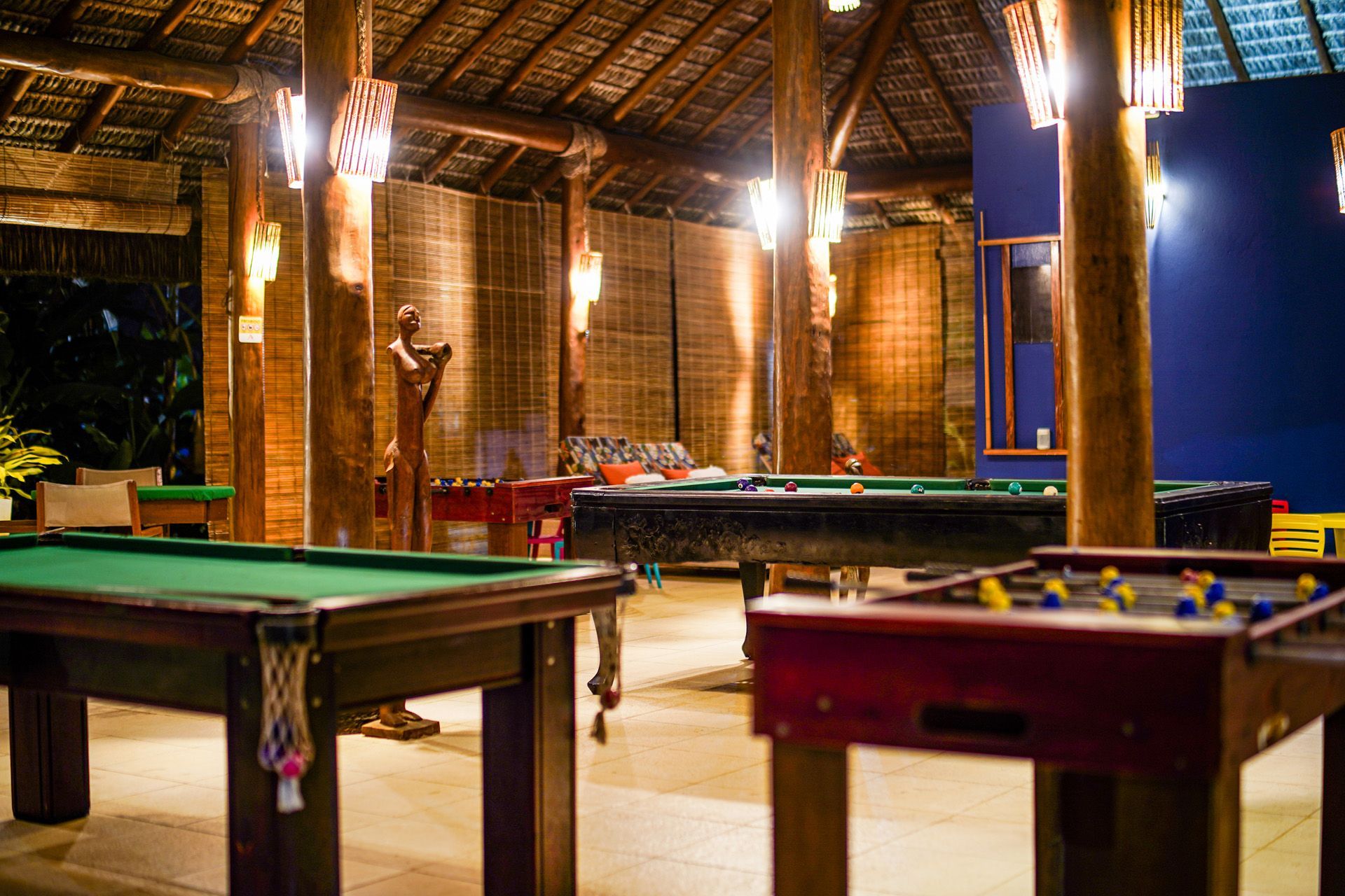 Sala de jogos com mesas de bilhar e matraquilhos, sob um telhado de palha com iluminação acolhedora.