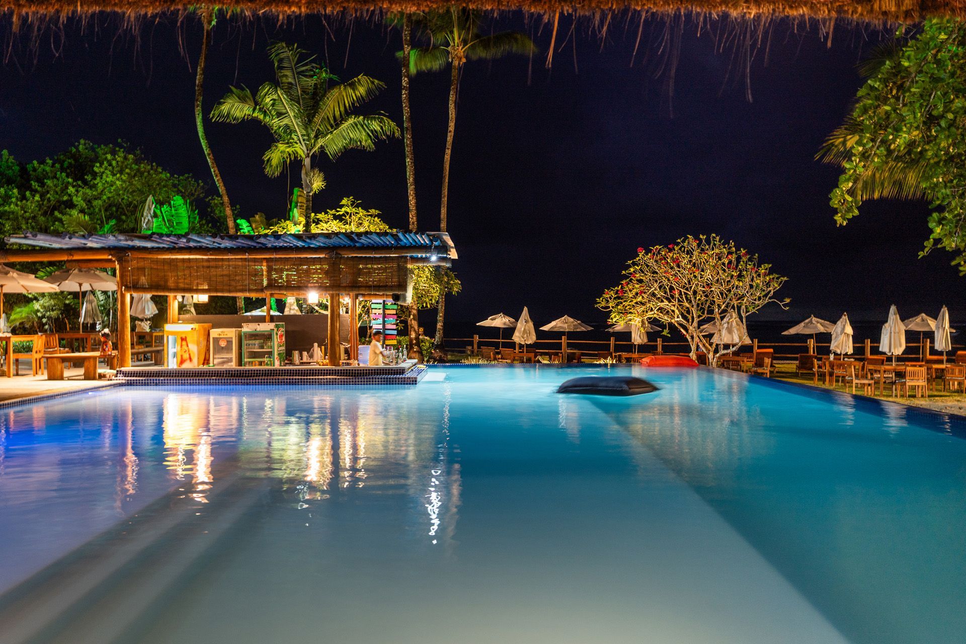 Duas mulheres em uma piscina, tomando champanhe em boias vermelhas sob um céu ensolarado em um resort.