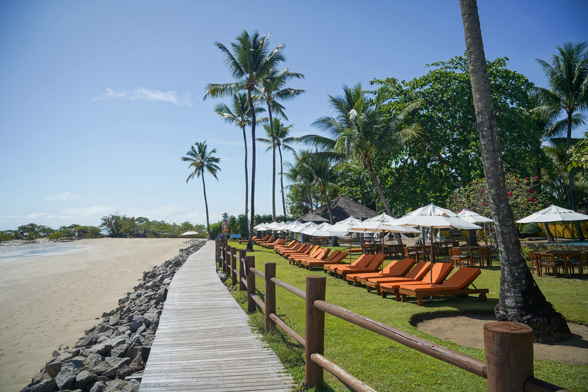 Passarela de madeira ao longo da praia com espreguiçadeiras, guarda-sóis e palmeiras sob um céu azul.