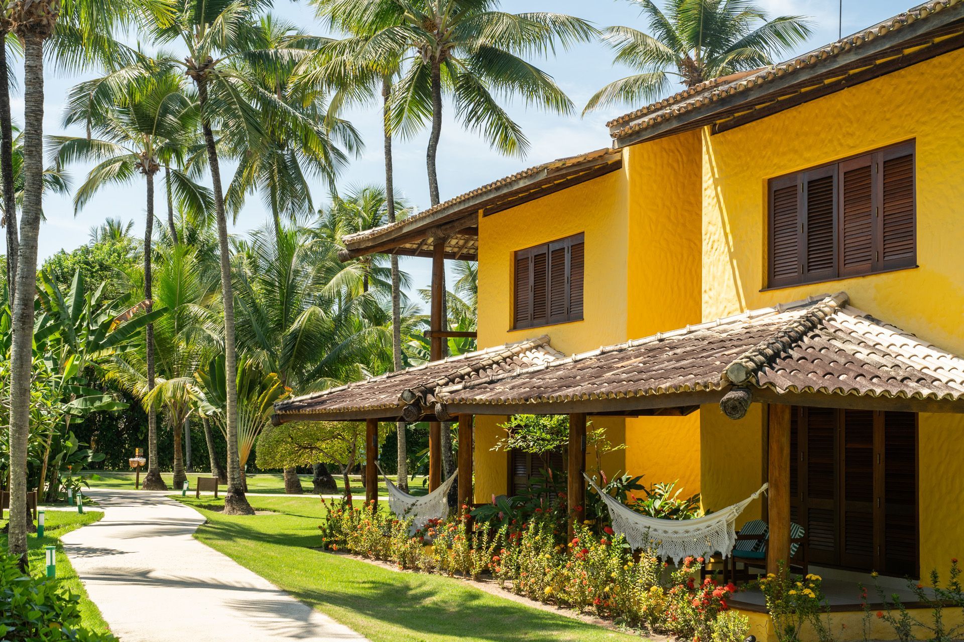 Prédio amarelo com telhado de telhas, varanda e redes; palmeiras e um caminho em um ambiente tropical.