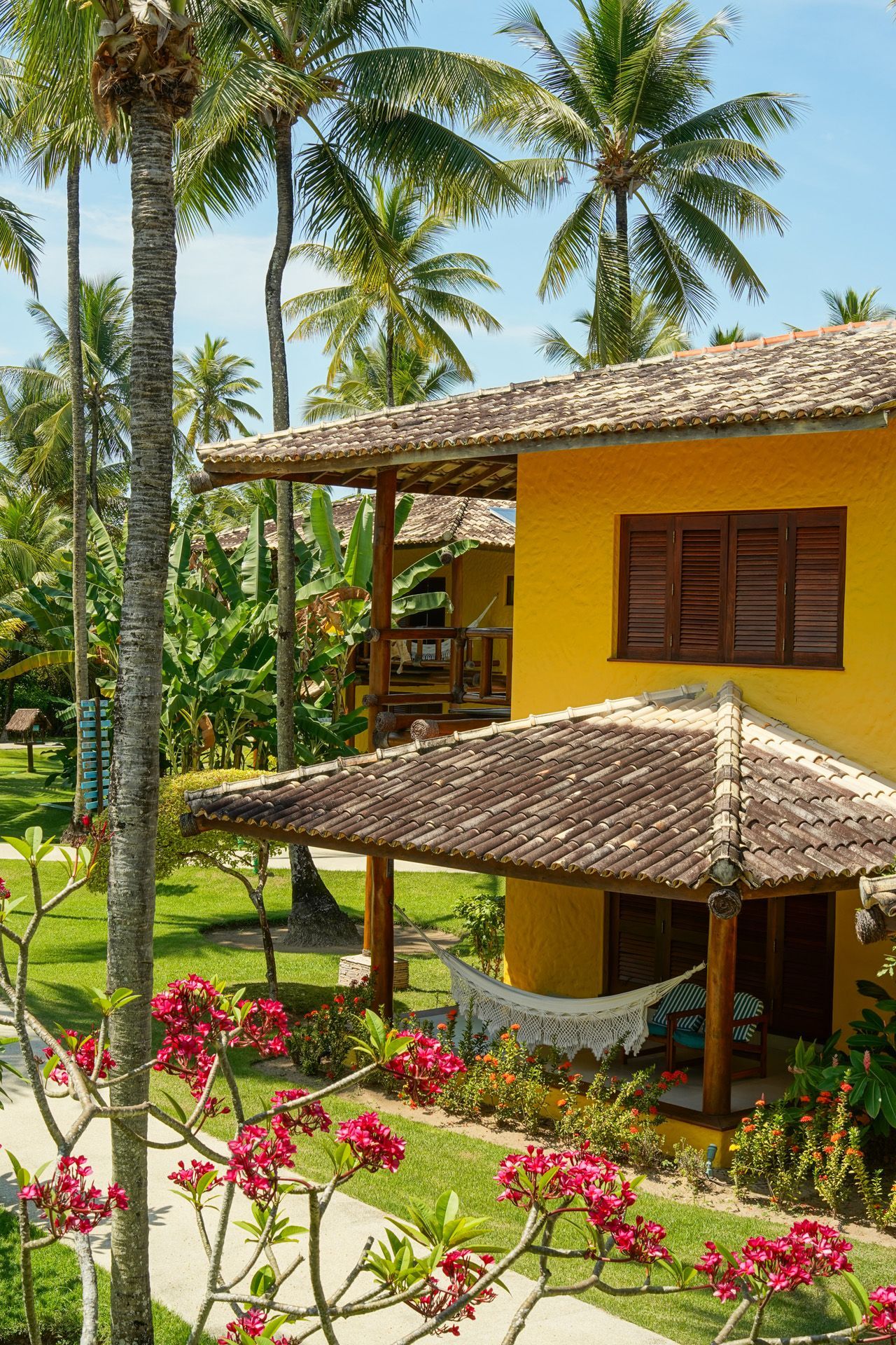 Casa tropical amarela com telhado de telha, varanda e rede. Palmeiras e gramado verde.