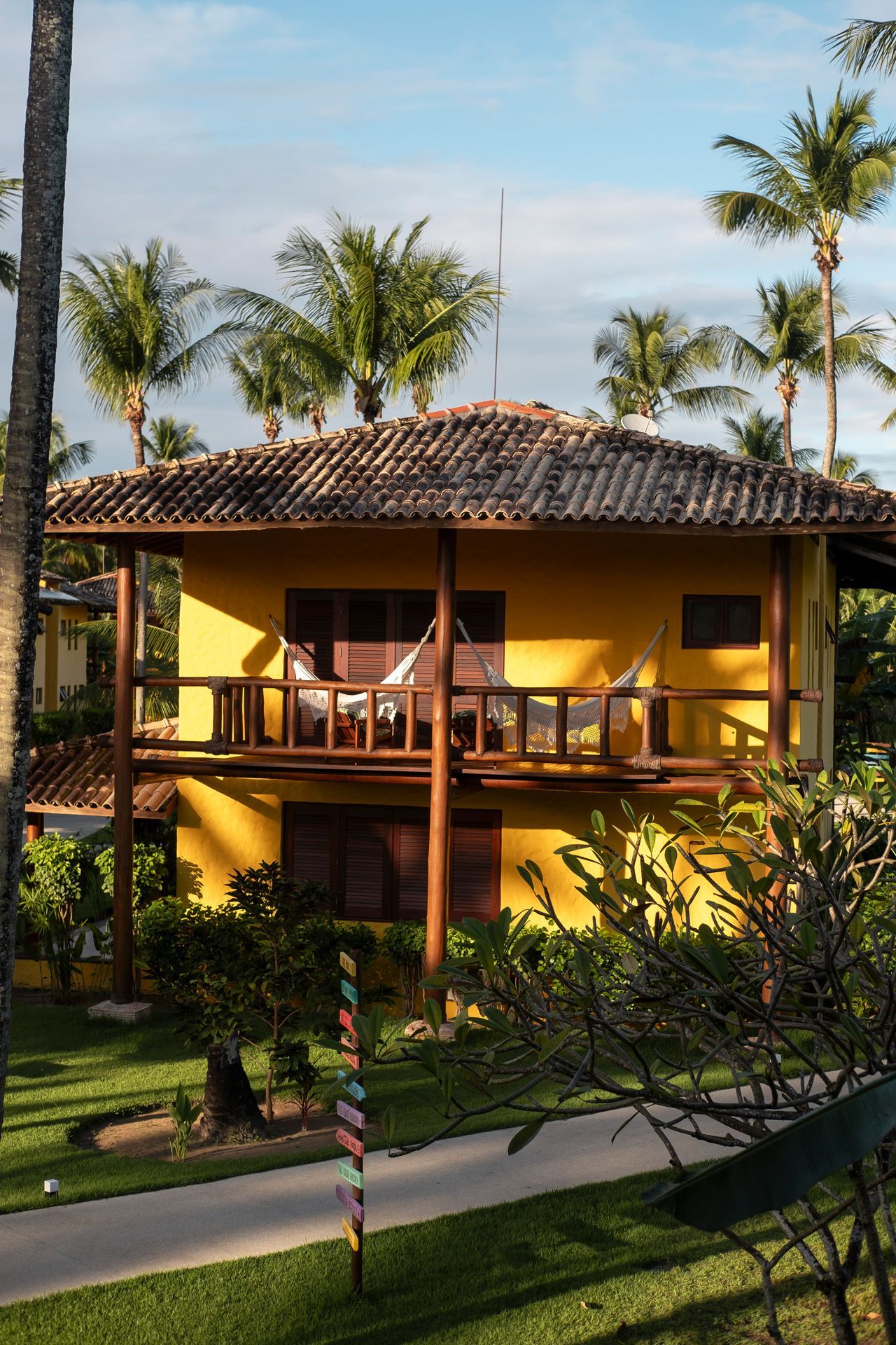 Prédio amarelo de dois andares com varanda, rede e telhado de telhas, situado entre palmeiras e grama verde.