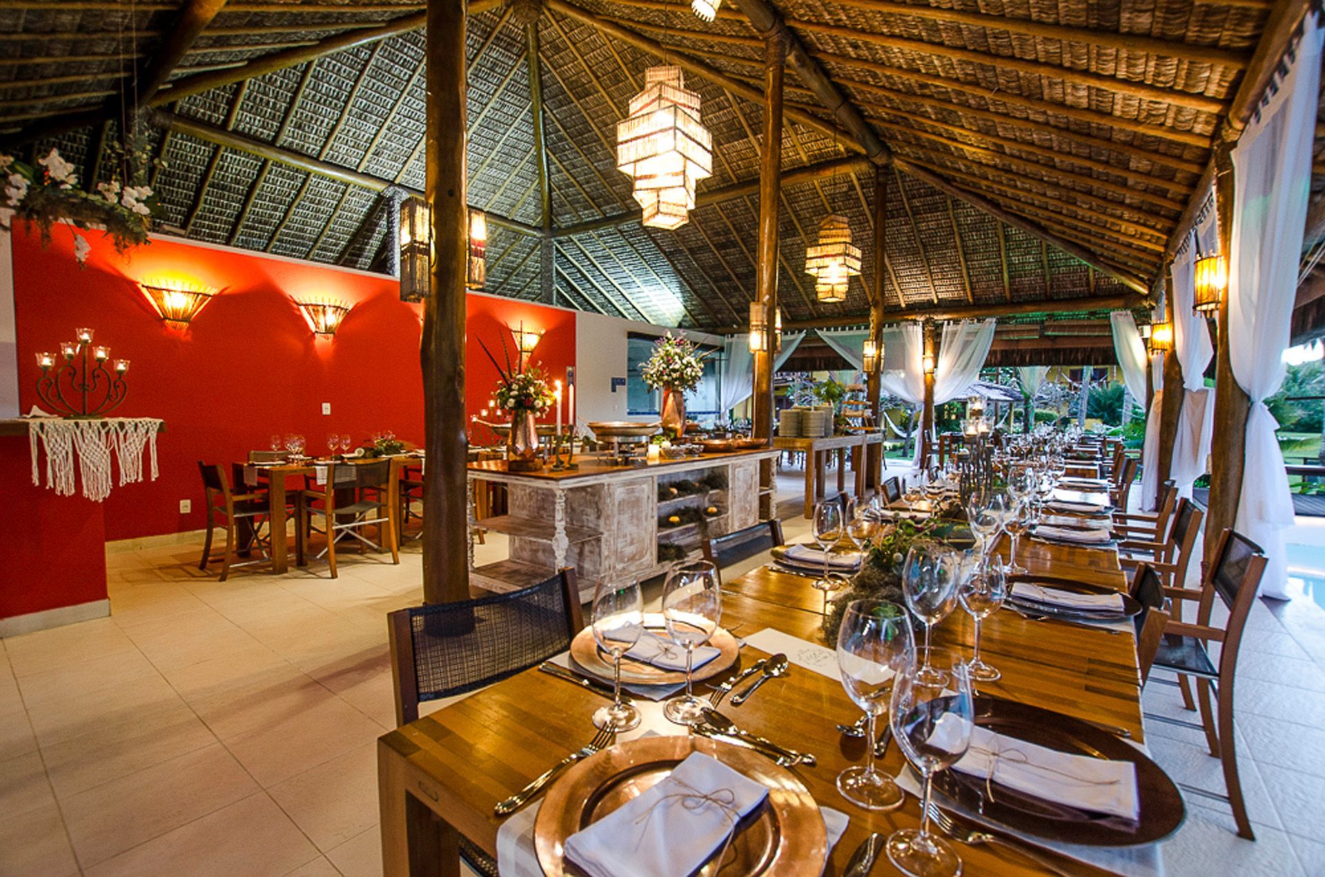 Área de refeições ao ar livre com mesas postas para uma refeição, sob uma estrutura de telhado de palha.