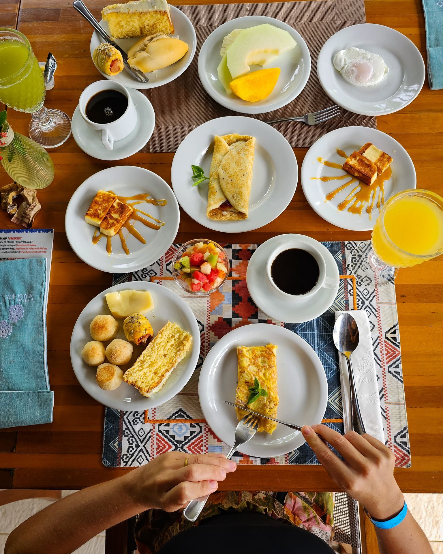 Mesa de madeira com uma mesa de café da manhã repleta de pratos e bebidas variadas. Uma pessoa está comendo, com os braços visíveis.