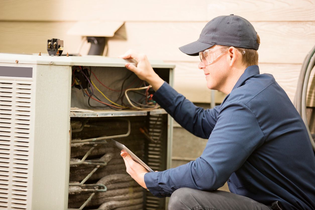 A technician in a blue uniform inspecting an open outdoor HVAC unit while holding a tablet.
