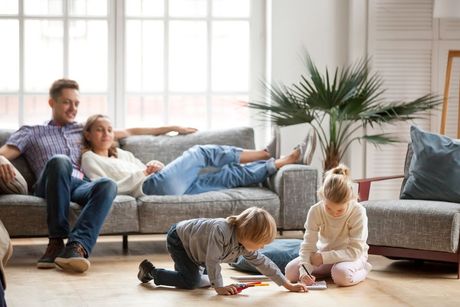 A family relaxes in a sunny living room as two children play on the floor and two adults lounge on a gray sofa.