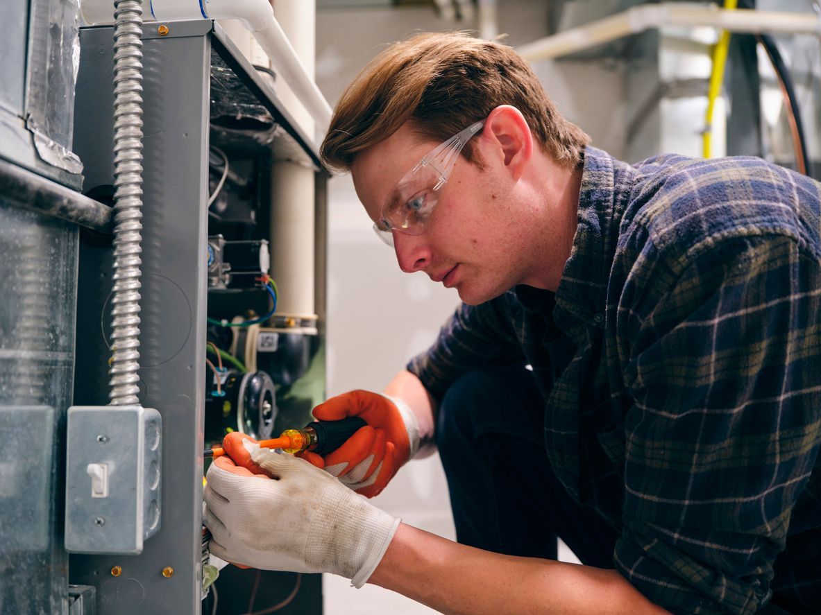 A technician wearing safety glasses and gloves uses a screwdriver to repair a furnace unit.