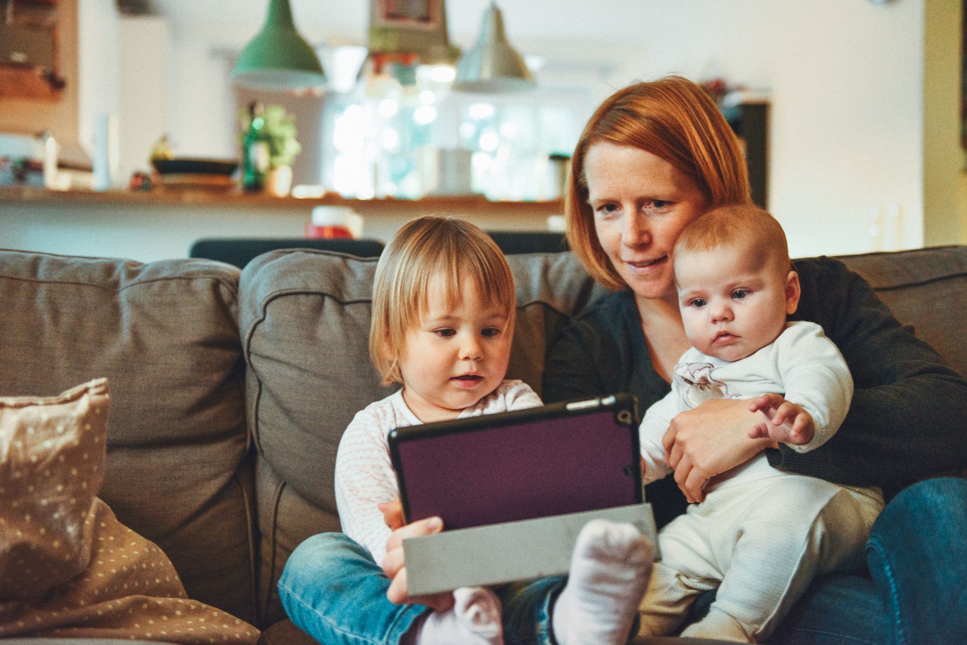 A parent sits on a couch with two children, all focused on a tablet held by the older child.