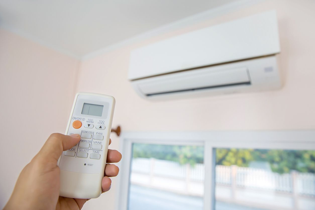 A hand holds a white remote control pointed toward a white wall-mounted air conditioner in a room.
