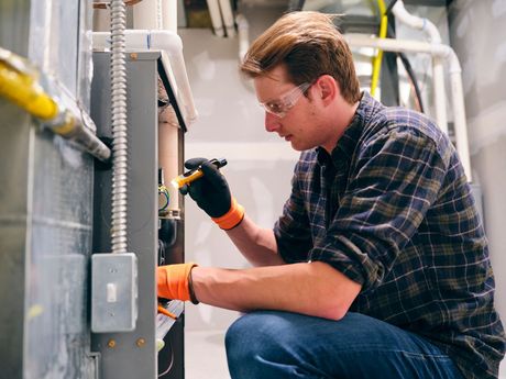 A technician in safety glasses and work gloves uses a flashlight to inspect the interior of a furnace.