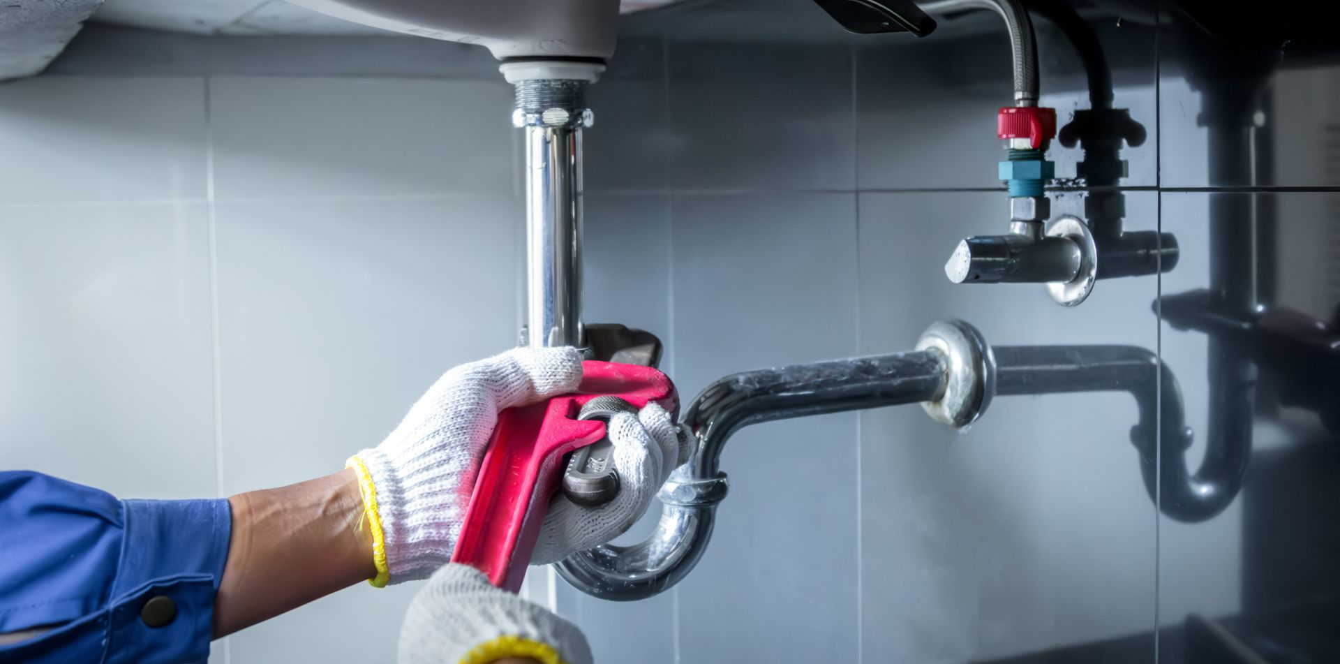 A worker wearing gloves uses a red wrench to tighten a metal pipe under a bathroom sink.