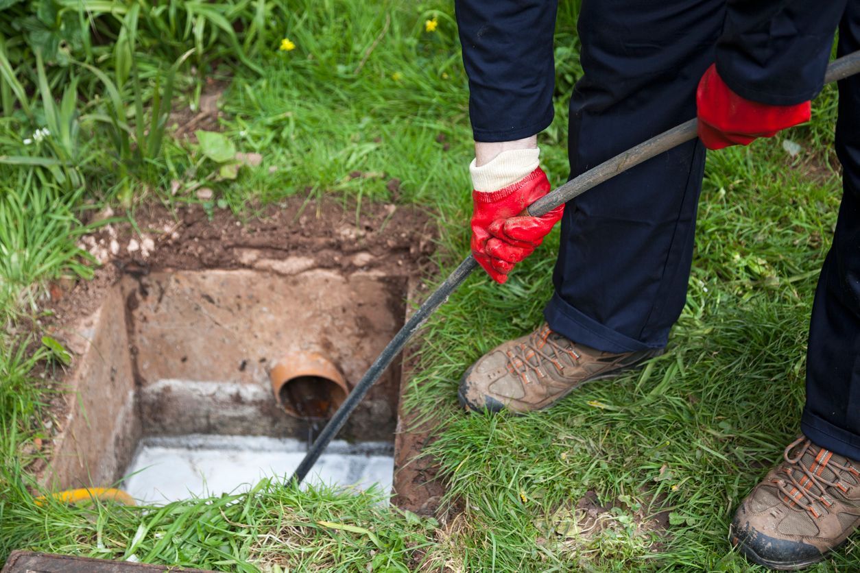 A worker wearing red gloves uses a drain snake to clear a residential sewer access point in a grassy yard.
